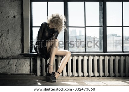 Tall model sitting on the windowsill in dark midi leather skirt and transparent blouse. Designer clothes. Artistic studio shot in industrial style interior.