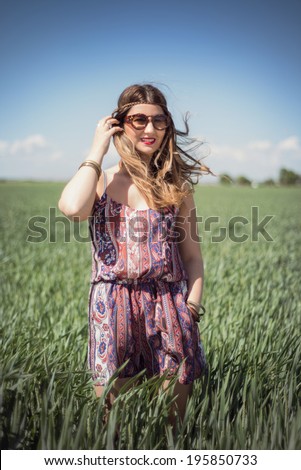 Beautiful fashion hippie woman in a wheat field wearing a boho jumpsuit romper and sunglasses, with the wind blowing in her blonde hair