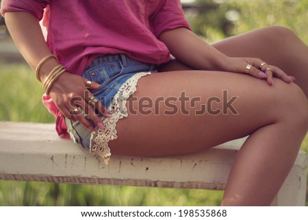 Sexy and attractive woman legs and hands, wearing sexy casual denim shorts with macrame, lace attachment a pink shirt and her hands full of gold fashion accessories rings bracelet