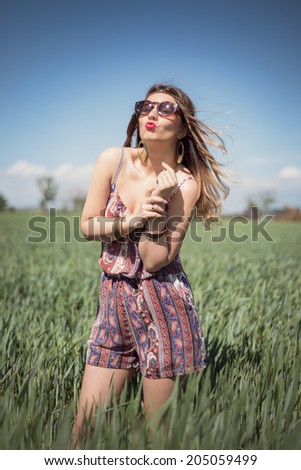 Beautiful fashion hippie woman in a wheat field wearing a boho jumpsuit romper and sunglasses, with the wind blowing in her blonde hair