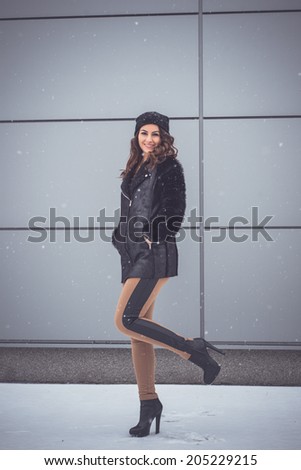 Woman posing on one foot in snow winter scene wearing a warm fur coat and sexy nude leggings with leather stripes on sides