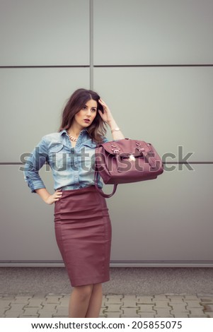 Beautiful business woman dressed in an office look, maroon pencil leather dress and denim shirt posing outdoor in an urban location