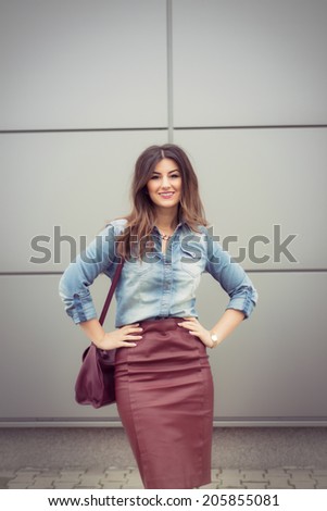 Beautiful business woman dressed in an office look, maroon pencil leather dress and denim shirt posing with a big smile and hands in her waist