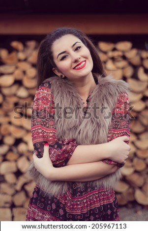 Portrait of Fashion beauty posing in front of a pile of wood wearing a beige fur vest, colorful dress hugging herself. Wood background