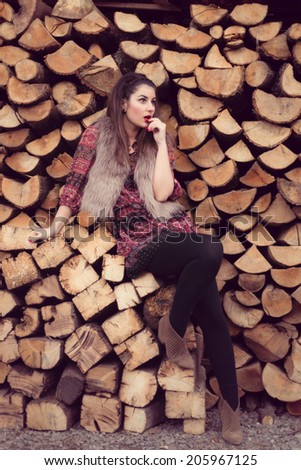Fashion beauty sitting on a pile of wood wearing a beige fur vest, colorful dress and black tights