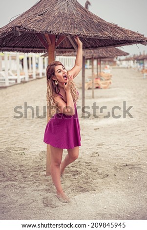 Fashion blonde happy at the sea posing shoe-less on sand under tiki patio umbrella dressed in a fuchsia pink dress holding hand at her mouth