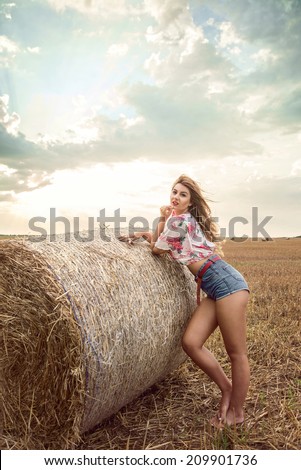 Beautiful woman fashion posing on wheat field at sunset in front of bales of straw, wearing sexy floral crop top and denim shorts. Incredible Cloudy sky