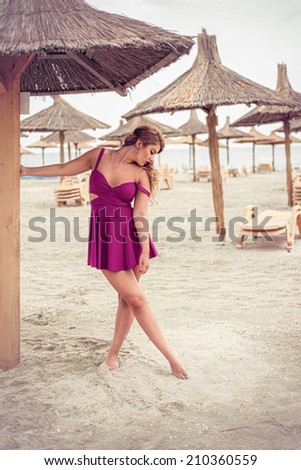 Fashion blonde happy at the sea posing sensual shoe-less on sand under tiki patio umbrella dressed in a fuchsia pink dress
