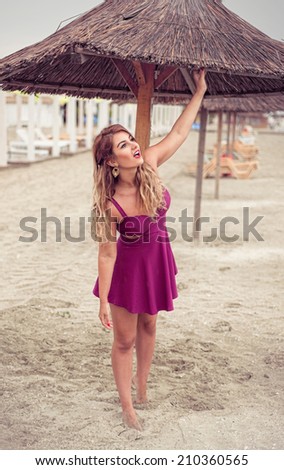Fashion blonde happy at the sea posing shoe-less on sand under tiki patio umbrella dressed in a fuchsia pink dress looking for rain