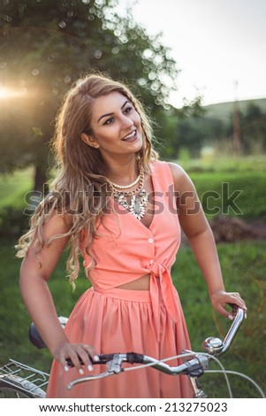 Pretty blonde riding her bicycle, wearing a sexy cropped top light pink dress. Sunset passing through the trees