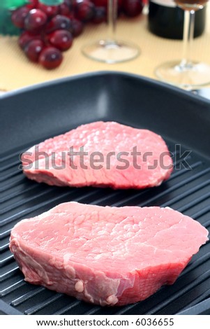 Two raw round eye steaks on a grilling pan with vegetables and wine in background