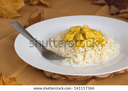 Small pieces of chicken cooked with onion and cream, curry, curcuma and ginger. Served on white plate with rice on natural brown background.