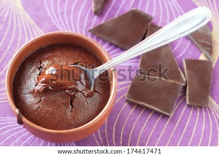 Traditional french chocolate fondant cake with melted heart in natural brown earthenware cup with a spoon. Natural blown and purple background with broken chocolate pieces.
