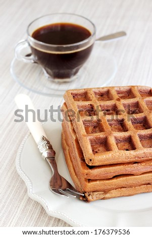 A bunch of chocolate gold brown waffles on light pastel vintage plate and natural napkin. Served with a transparent cup of coffee.