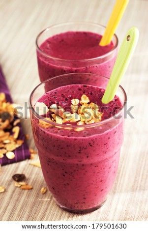 Smoothie made with frozen red berries, banana and milk in transparent glass. Green spoon. Decorated with muesli preparation. Natural brown napkin and violet napkin on the background.