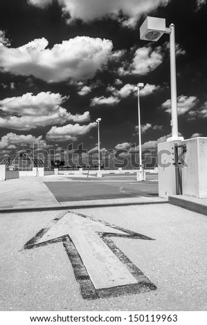 Black and white image of an arrow and street lamp under a partly cloudy sky, on top of a parking garage in Towson, Marylnd.