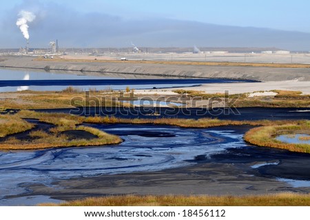 Alternating stripes of bitumen, water, sand and grass at a mine\'s tailings pond, where fine waste gradually settles. Also one oil barrel visible.