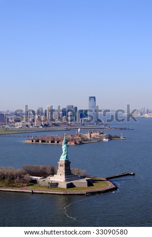 Aerial of the Statue of Liberty in the New York City harbor on a clear spring day. Behind Liberty Island is Ellis Island and then skyscrapers of Jersey City.