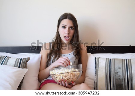Latina woman watching a movie in her bed, while eating popcorn. Surprised expression.
