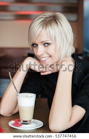 Beautiful young blond woman seated at a table in a cafeteria enjoying a large glass of frothy cappuccino coffee resting her chin on her hands and smiling at the camera