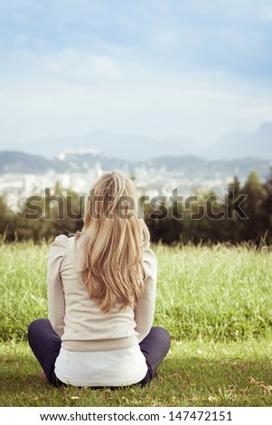 Woman enjoying a view of the town sitting cross legged in a green field with her back to the camera