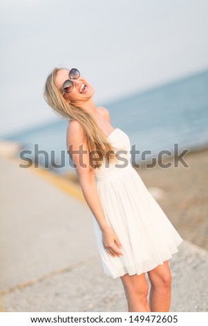 Pretty woman wearing sunglasses enjoying the summer at the beach standing on a walkway laughing happily in the sunshine