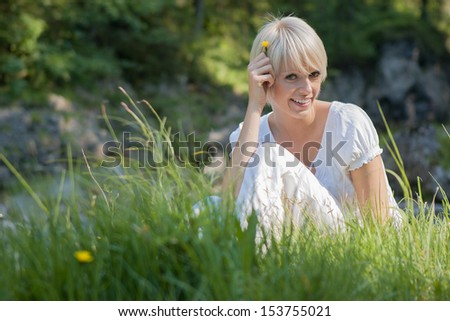 Beautiful young blond woman in nature sitting in long fresh green grass playfully holding a dandelion to her hair and smiling