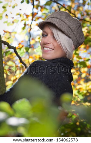 Beautiful trendy young woman wearing a stylish cloth cap turning to look at the camera with a smile standing in autumn trees
