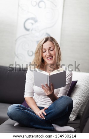 Attractive young blond woman relaxing at home reading a book sitting cross legged on a sofa in the living room