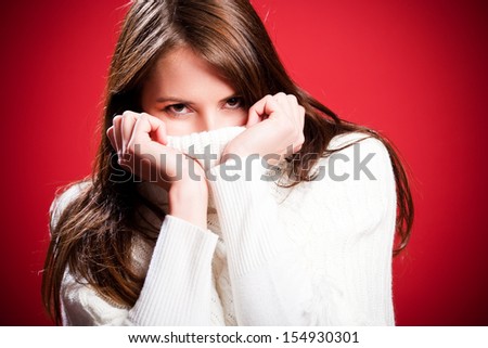 Woman snuggled down inside her warm winter top pulling the collar up over her face so that only her eyes are visible watching the camera, over a festive red background with vignetting