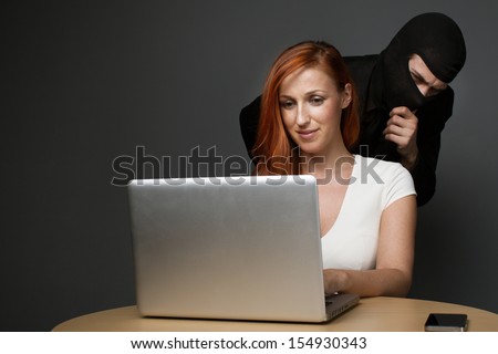 Man in a balaclava furtively watching an unsuspecting female office worker working on her laptop computer while corporate spying, stealing personal or business information or employee monitoring