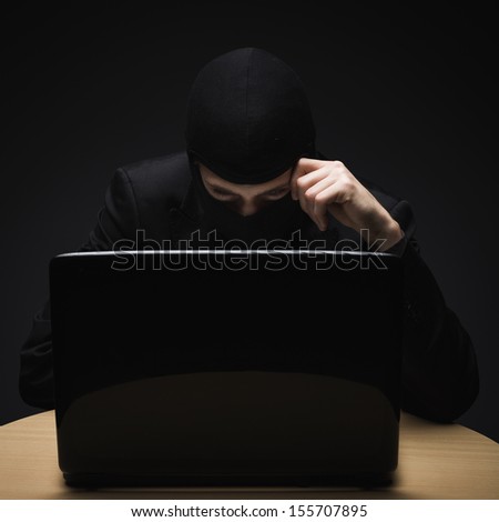 Cyber crime as a hacker disguised in a balaclava sits in the darkness behind a laptop computer stealing personal or business information