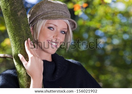 Beautiful young woman with a beaming smile standing with her arm around a tree trunk in woodland enjoying nature and the outdoors