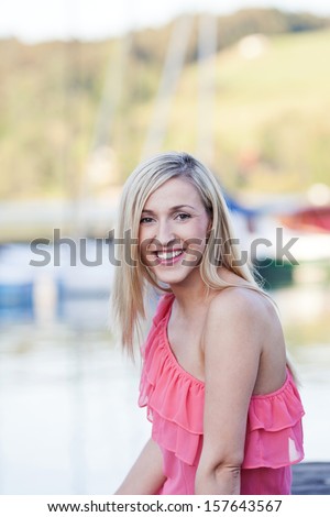 Pretty young blond woman outdoors in a trendy pink summer top relaxing at the seaside or lake on a summer vacation