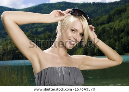 Stunning trendy blond woman in an off the shoulder summer dress and her sunglasses pushed to the top of her head standing with her arms raised smiling at the camera outdoors in the mountains