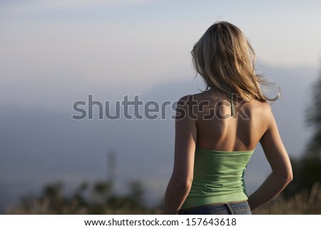Solitary young woman with long blond hair standing with her back to the camera looking out over a misty mountain view deep in contemplation in a depiction of Loneliness