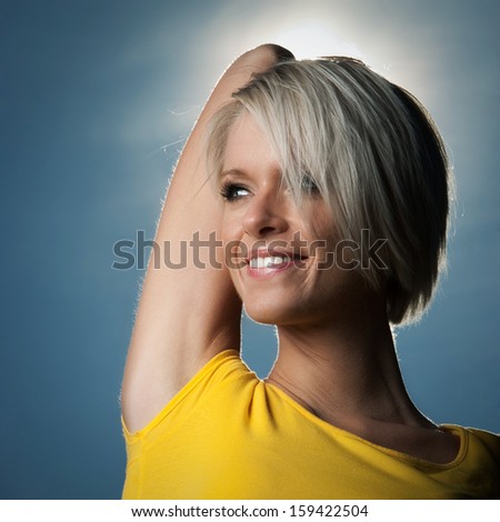 Close up head and shoulders portrait of a smiling beautiful young woman with a short modern blond hairstyle standing backlit by a sunburst with her arm raised behind her head