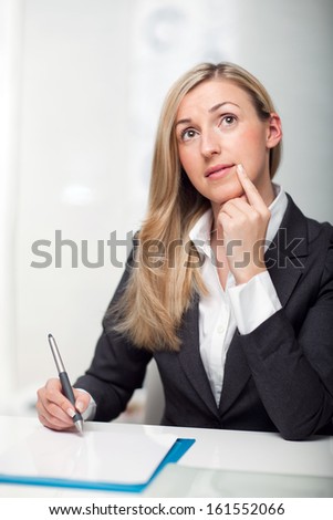 Thoughtful young businesswoman sitting at her desk writing notes looking up in contemplation with her finger to her chin