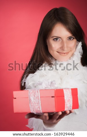 Beautiful stylish woman in a glamorous feathery outfit holding out a red gift box for Christmas or Valentines with a lovely smile