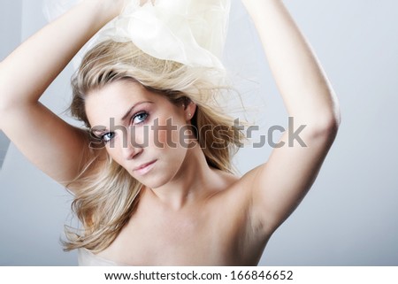 Beautiful graceful blond woman with her arms raised in the air and long hair tossed over her bare shoulder looking at the camera with a serious expression, studio head and shoulders portrait on grey