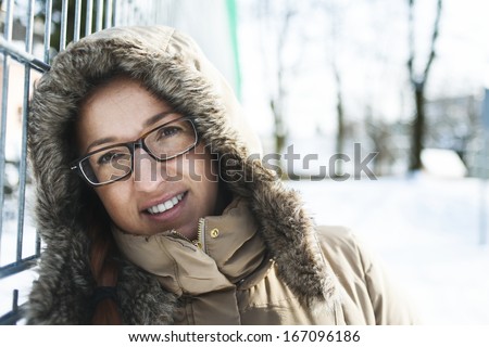 Close up head and shoulders portrait of a woman in winter fashion wearing glasses and a hooded jacket trimmed with fur standing outdoors, in a wintry landscape