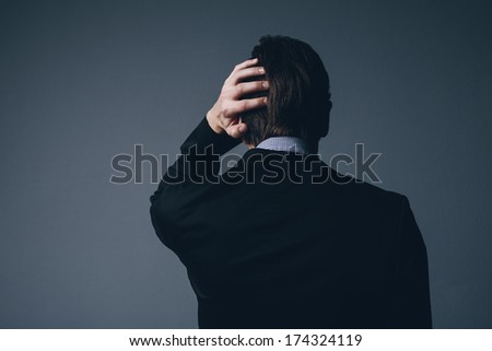 View from behind of a stylish businessman standing with his hand to his head as he scratches it in perplexity or as he nurses a throbbing headache on a dark grey background