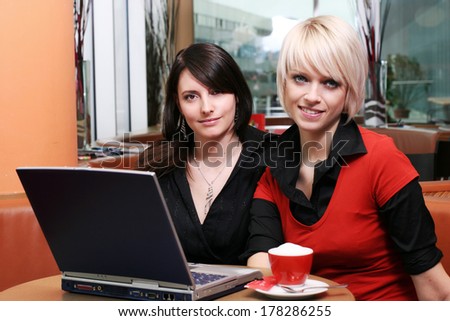 Two beautiful women friends meeting over coffee in a restaurant sit at a table together sharing a laptop computer and looking at the camera with friendly smiles