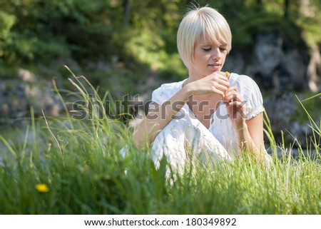 Low angle view through green grass of a beautiful young blond woman in a white summer blouse sitting playing with meadow flowers in the sunshine