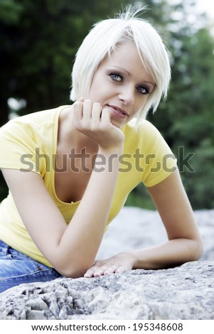 Beautiful friendly young blond woman with a trendy short hairstyle reclining on a rock resting her chin on her hand smiling at the camera