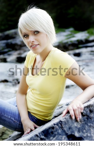 Trendy beautiful young blond woman sitting outdoors on a rock enjoying the peace and quiet of nature turning to look at the camera with a smile