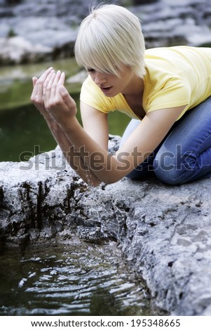 Beautiful young woman cupping water from a rock pool kneeling down on the rocky edge letting the cool water fall in droplets from her hands