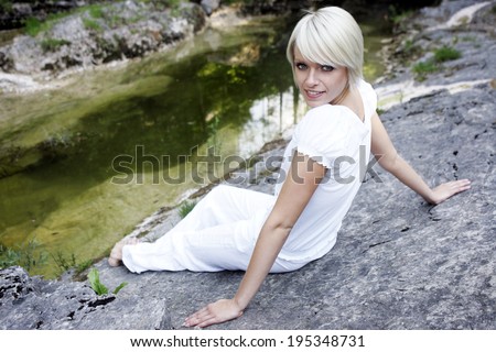 Beautiful young blond woman in fresh white clothing relaxing on a rock in the summer sunshine turing her head to smile up at the camera