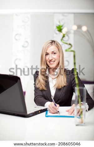 Beautiful businesswoman in a modern office sitting at her desk in front of a laptop computer writing notes on a pad and looking at the camera with a smile