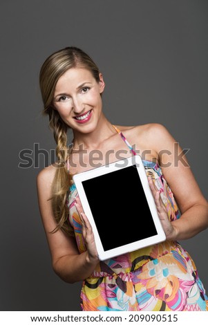 Pretty woman with her long blond hair in a plait wearing a floral summer dress holding a tablet-pc with the blank screen towards the camera